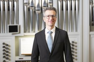 Wolfgang Kogert Portrait 2025 im Anzug vor der Orgel der Wiener Hofburgkapelle